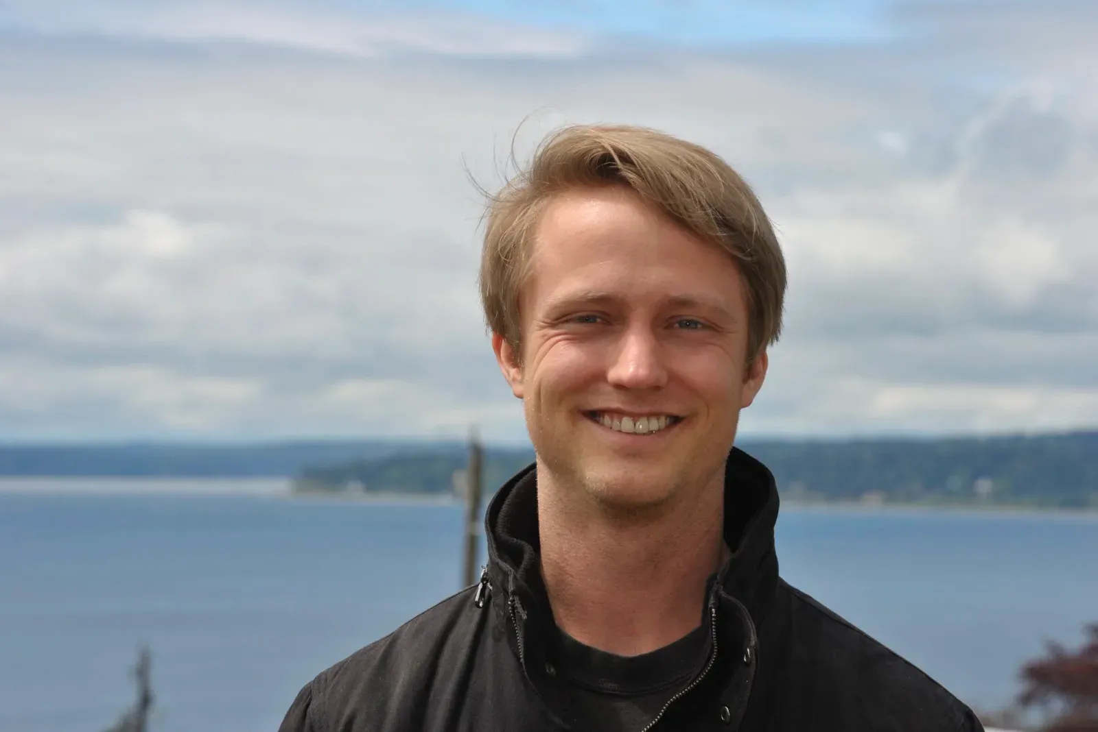 Jake Douglass smiling outdoors, with water and a wooded shoreline behind him.